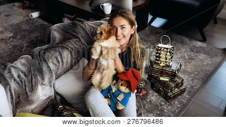 Smiling Lady With Long Blonde Hair Sitting On Comfortable Sofa With Pillows And Holding Little Doggy