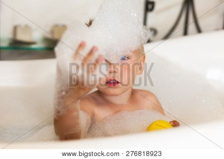 Little Caucasian Boy With Blue Eyes In The Bath Pulls One Hand Towards, In Bubbles Of Foam, With Yel