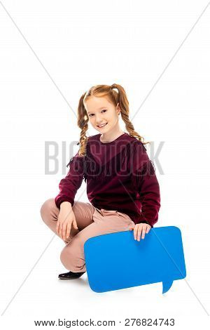 Smiling Schoolgirl Sitting On Hunkers, Holding Speech Bubble And Looking At Camera Isolated On White