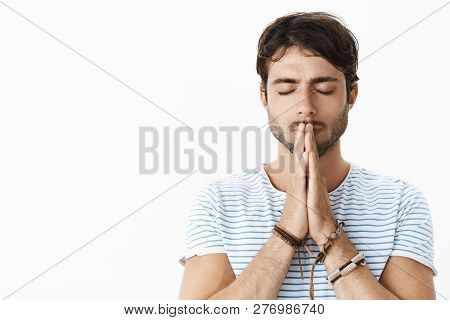 Focused Young Hopeful Hispanic Guy With Beard In Striped T-shirt Holding Hands In Pray Over Lips Sta