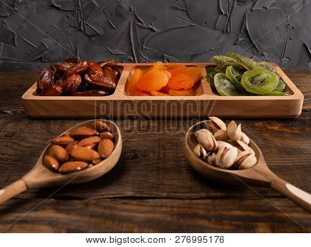 Dates, Dried Apricots And Kiwis In A Compartmental Dish And Nuts In A Wooden Spoon On A Dark Wooden 