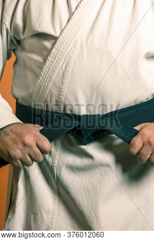 A Man Is Tying A Black Belt On A White Kimon On A Background Of Ora.