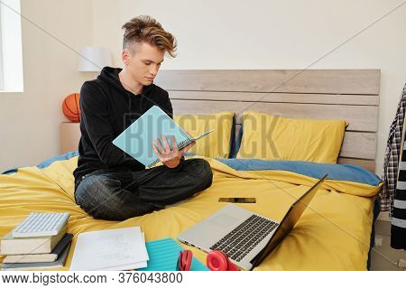 Software Engineering Student Sitting On Bed With Books, Laptop And Textbooks Around And Writing In N