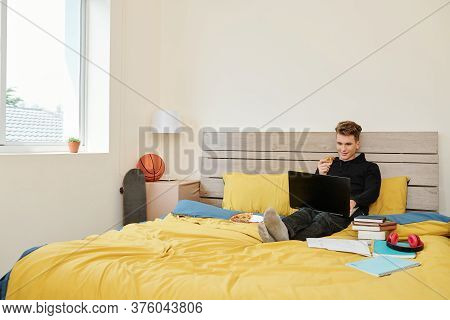 Positive High School Student Sitting On Bed, Eating Pizza And Watching Online Webinar When Staying H