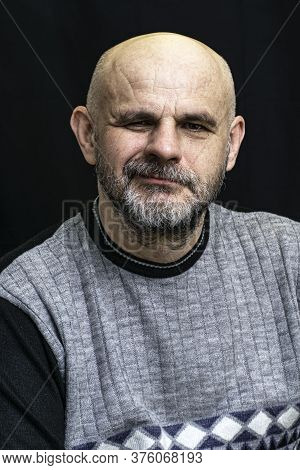 Middle-aged Bald Bearded Man Grins Contemptuously Against The Dark Background