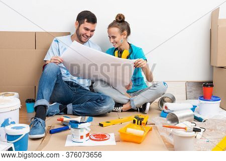 Happy Couple Sitting On Floor With Construction Blueprint. Home Remodeling And House Interior Redesi