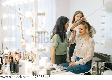 Two Professional Female Make-up Artist Doing Makeover In Lighting Dressing Room, View From Afar. Con