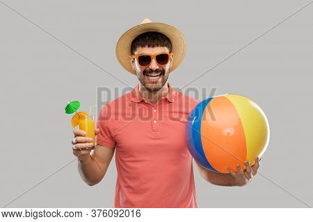 summer, vacation and people concept - happy smiling young man in sunglasses and straw hat with orange juice cocktail and inflatable beach ball over grey background