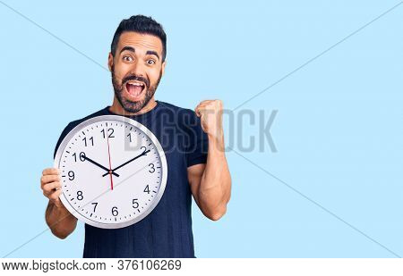 Young hispanic man holding big clock screaming proud, celebrating victory and success very excited with raised arms 