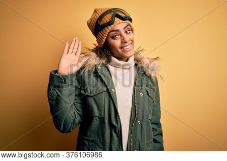 Young african american afro skier girl wearing snow sportswear and ski goggles Waiving saying hello happy and smiling, friendly welcome gesture