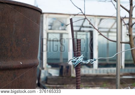 Old Tank And Pin With A Metal Wire, Against The Background Of A Greenhouse And A Young Apple Tree
