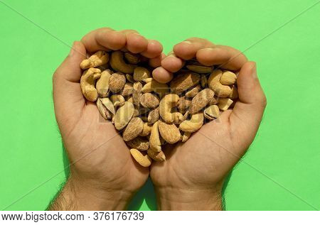 Mixed Nuts In Man's Hands Forming Heart Shape On Green Background. Top View. Salted And Spicy Pistac