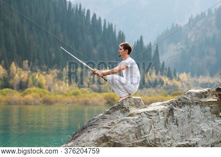 A Barefoot Man Sits On A Stone And Holds A Sword In His Hands. Preparing For The Battle. Man Health 