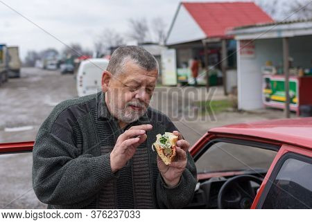 Senior Driver Holding Street Food (lyulya Kebab In Lavash) Near His Car While Standing On A Back Roa