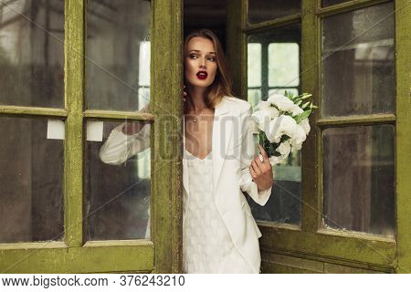 Young Gorgeous Woman In White Jacket And Dress Holding Little Bouquet Of Flowers In Hand Dreamily Lo