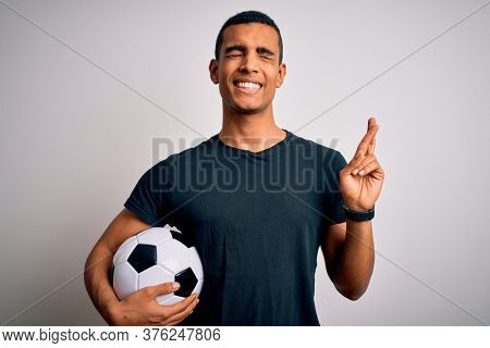 Handsome african american man playing footbal holding soccer ball over white background gesturing finger crossed smiling with hope and eyes closed. Luck and superstitious concept.