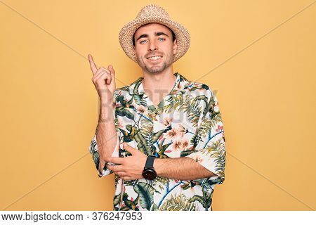 Young handsome man with blue eyes on vacation wearing summer florar shirt and hat with a big smile on face, pointing with hand and finger to the side looking at the camera.