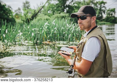 Young Fisherman Fishing On Lake Or River. Busy Cool And Serious Guy In Singlasses Holding Fish Plast