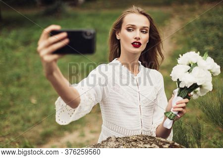 Young Beautiful Woman In White Dress Holding Little Bouquet Of Flowers In Hand While Thoughtfully Ta