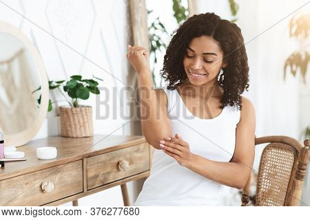 Dry Elbows Treatment. Smiling Black Woman Applying Moisturising Cream At Home, Healing Cracked Skin