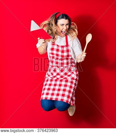 Young beautiful blonde cook woman wearing apron smiling happy. Jumping with smile on face holding recipe book and spoon over isolated red background