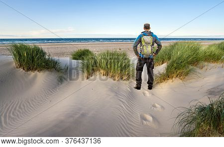 Man With Backpack Hiking In Beautiful Windy Coastal Dune Marram Grass Towards Beach Of North Sea In 