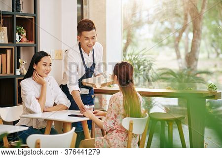 Young Smiling Waiter Serving Water To Pretty Young Women Sitting At Cafe Table