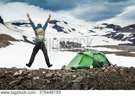 Hiker Man With Backpack And Tent Jumping With Outstretched Arms. Camping In Snow Mountain Glaciers A
