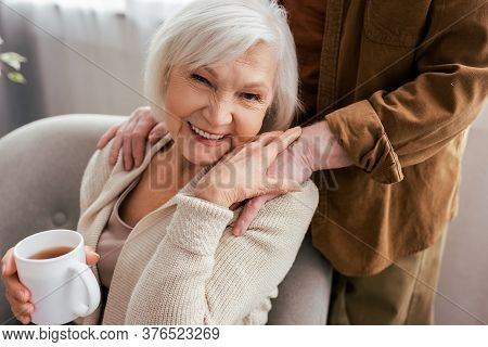 Cropped View Of Man Touching Shoulders Of Cheerful Senior Wife Holding Cup Of Tea And Smiling At Cam