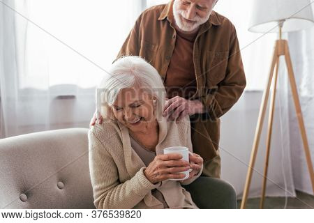 Cheerful Senior Man Touching Shoulders Of Happy Wife Sitting In Armchair With Cup Of Tea