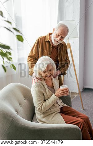 Smiling Senior Man Touching Shoulders Of Happy Wife Sitting In Armchair With Cup Of Tea