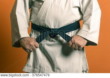 A Man Is Tying A Black Belt On A White Kimon On A Background Of Ora.