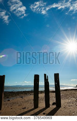 Old Pier Posts In Silhouette Cast Shadow On Sand At Low Tide With Lens Flare From Starburst Sun.