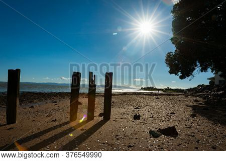 Old Pier Posts In Silhouette Cast Shadow On Sand At Low Tide With Lens Flare From Starburst Sun.