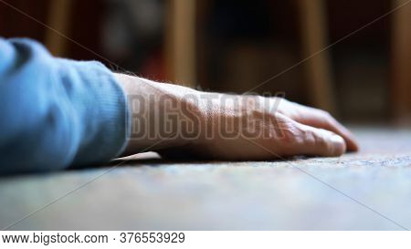 Aged Person In Blue Pullover Hand Lies On Floor Against Blurry Brown Wooden Furniture Under Bright S
