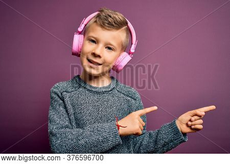 Young little caucasian kid wearing headphones listening to music over purple background smiling and looking at the camera pointing with two hands and fingers to the side.