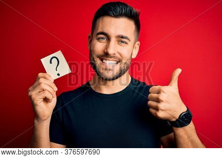 Young handsome man holding paper with question mark symbol over red background happy with big smile doing ok sign, thumb up with fingers, excellent sign