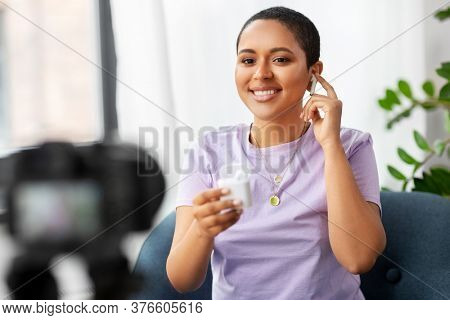blogging, videoblog and people concept - happy smiling african american female video blogger with camera and wireless earphones videoblogging at home