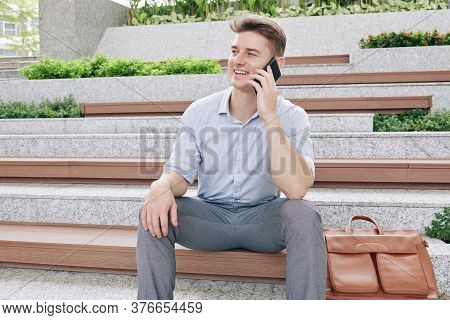 Positive Young Entreprenuer Sitting On Wooden Bench Outdoors And Talking On Phone With Colleague