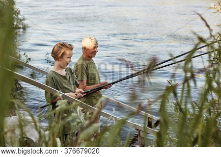 Fishing By Lake, Grandson And Grandfather Near River, Holding Fishing Rods In Hands, Look Excited An