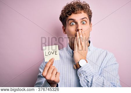 Young blond romantic man with curly hair holdiing paper with love message for valentines day cover mouth with hand shocked with shame for mistake, expression of fear, scared in silence, secret concept