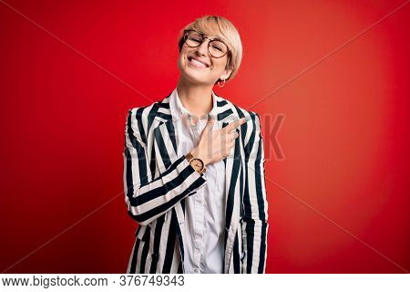 Blonde business woman with short hair wearing glasses and striped jacket over red background cheerful with a smile of face pointing with hand and finger up to the side with happy and natural expressio