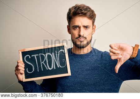 Young man holding blackboard with strong message standing over isolated white background with angry face, negative sign showing dislike with thumbs down, rejection concept