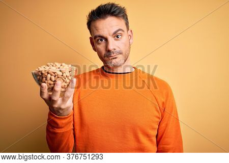 Young handsome man holding bowl with peanuts standing over isolated yellow background with a confident expression on smart face thinking serious