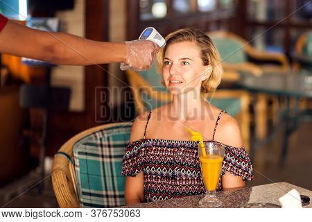A Worker Measuring Temperature Of Woman Visitior Inside  Of The Cafe. Life During Coronavirus Time