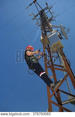 Tehnician In Red Helmet And Safety Belt Make Radio Call On The Electric Pole