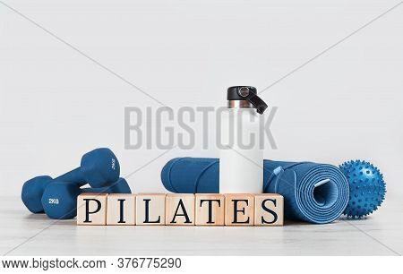 Pilates Text. Rolled Blue Yoga Mat And Excersise Resistance Band Dumbbells On Grey Wooden Surface. G