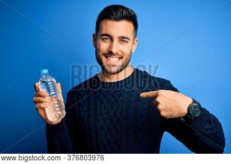 Young handsome man drinking bottle of water to refeshment over blue background with surprise face pointing finger to himself