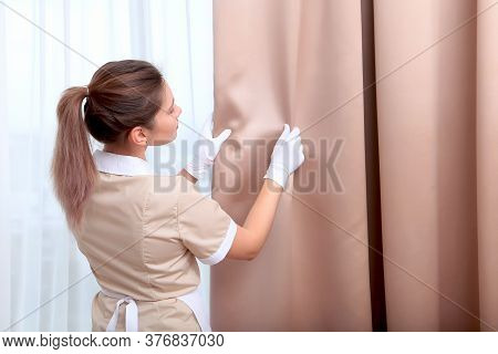 A Uniformed Maid Adjusts The Curtains In A Hotel Room. The Concept Of The Hotel Business. Cleaning I