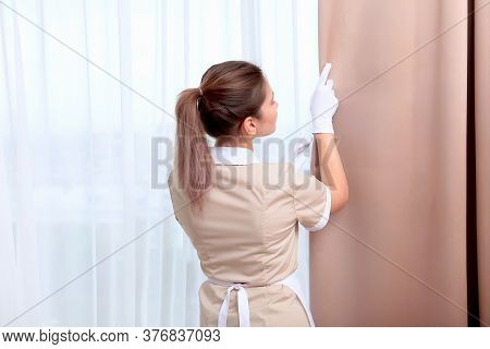 A Uniformed Maid Adjusts The Curtains In A Hotel Room Or House. The Concept Of The Hotel Business. C